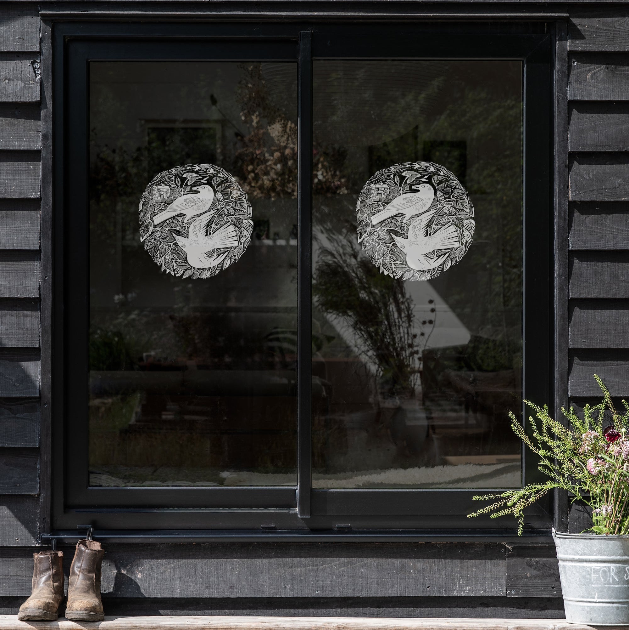 Two circular white blackbird window decals by Angela Harding applied to a black-framed sliding glass door on a dark gray wood-paneled wall. 